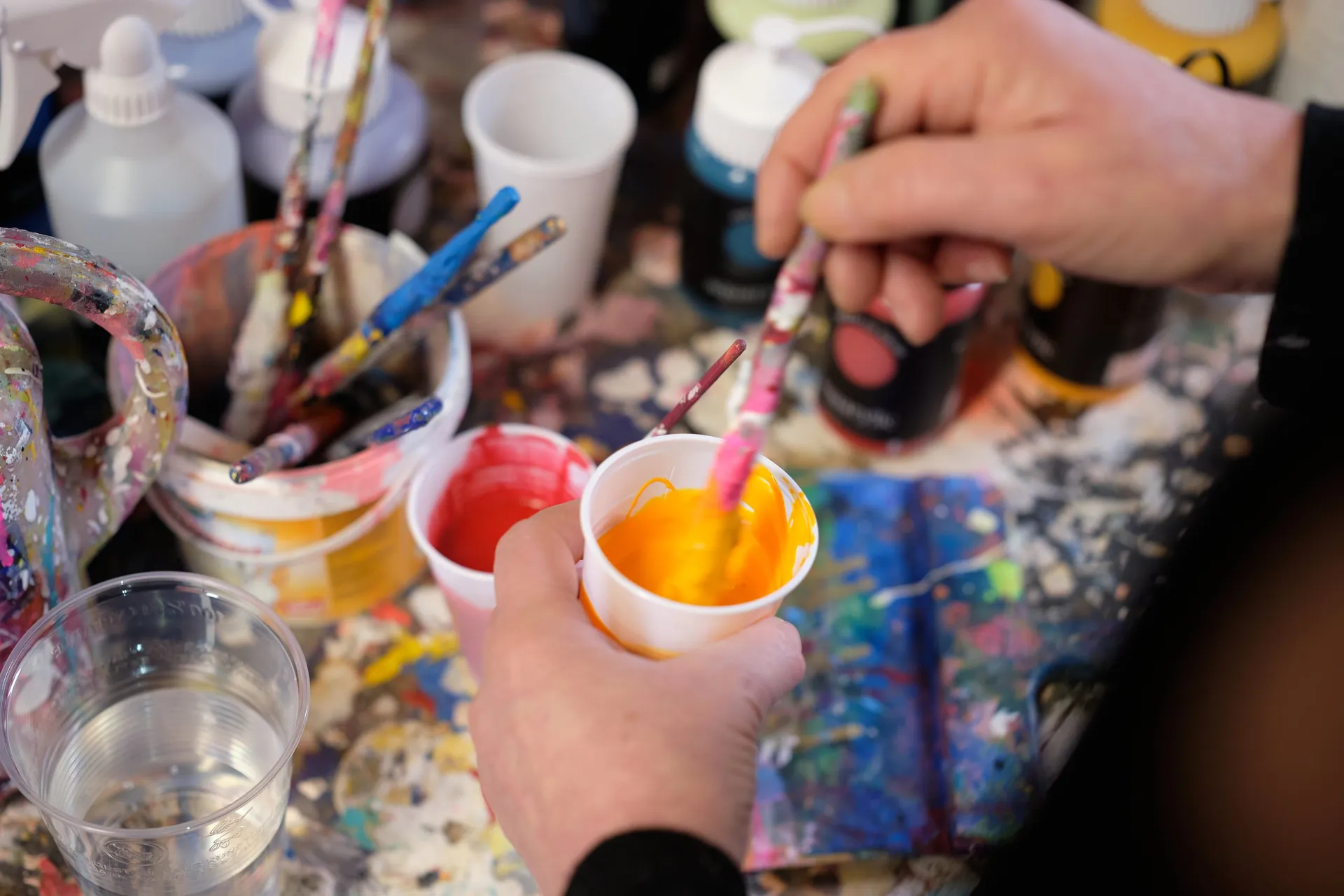 Eine Hand taucht einen Pinsel in einen Becher mit oranger Farbe, auf dem Tisch weitere Becher und Pinsel.