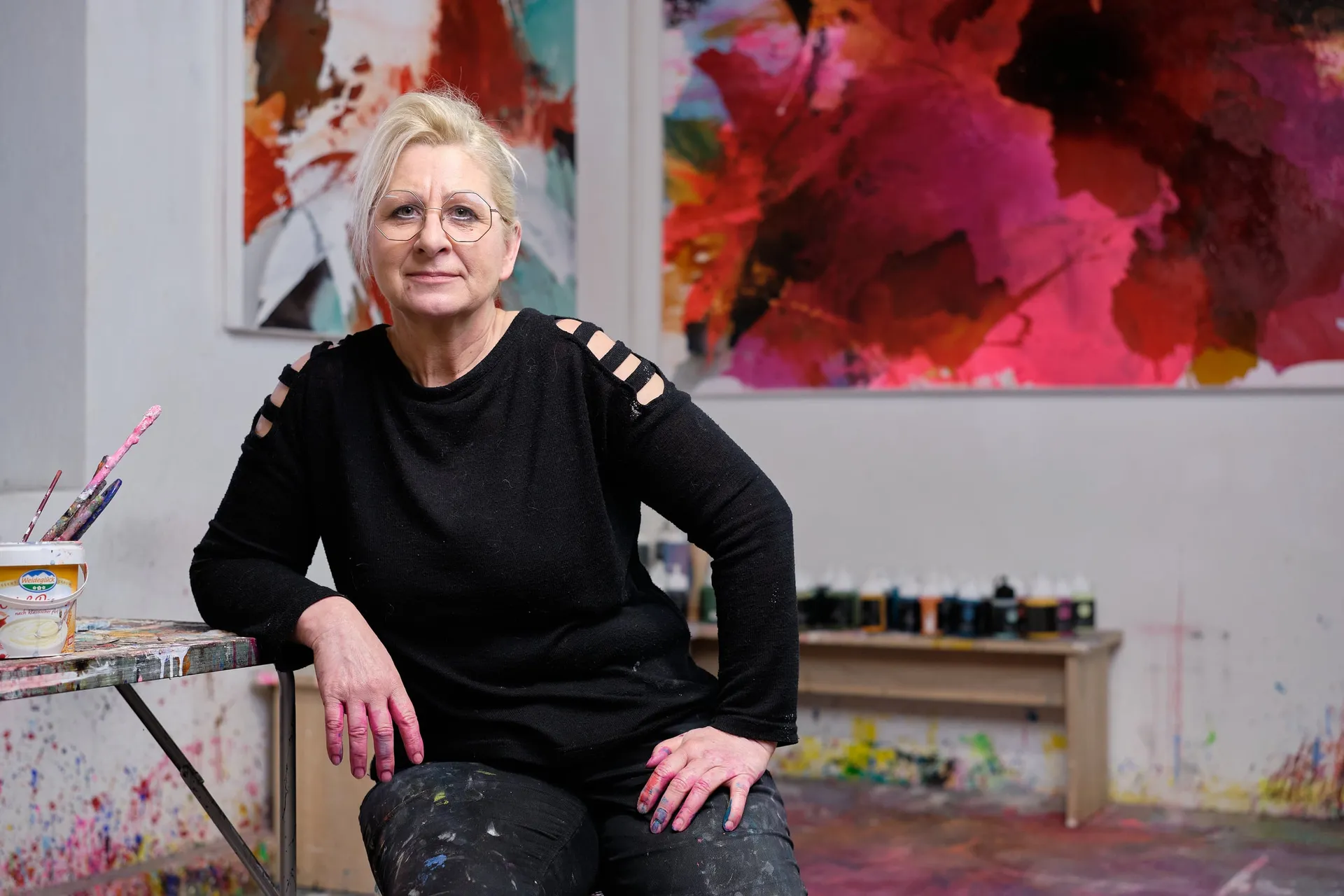 Eine Frau in schwarzem Pullover sitzt im Atelier vor großen, rot leuchtenden Gemälden.