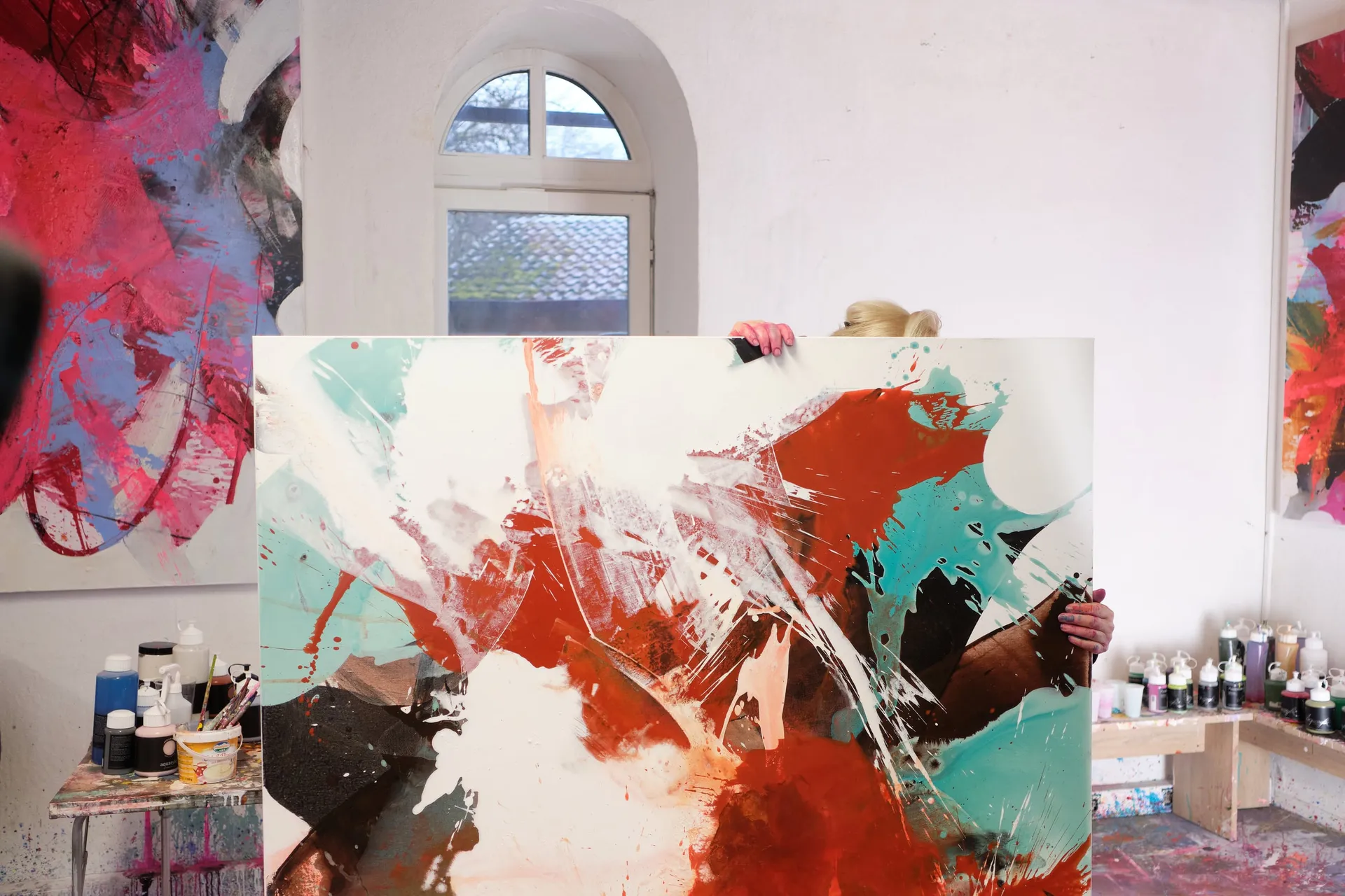 Eine Person hält im Atelier eine große Leinwand mit roten und türkisen Farbflächen vor sich.