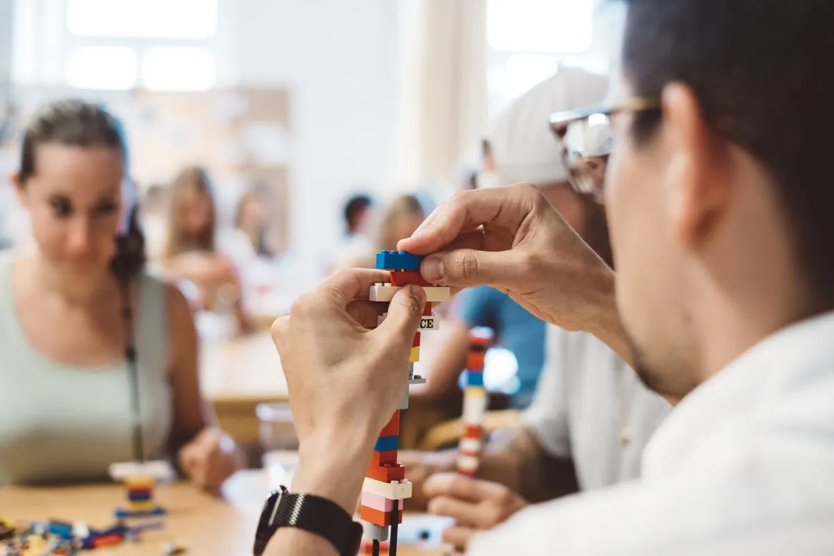 Mehrere Teilnehmende bauen am Tisch Figuren aus Lego-Steinen, im Hintergrund weitere Personen.