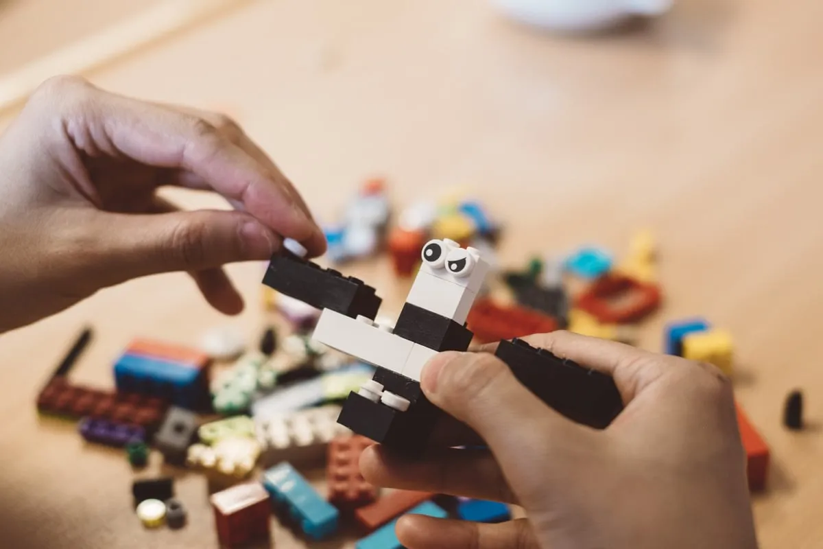 Hände setzen eine schwarz-weiße Figur mit Wackelaugen aus Lego-Steinen zusammen, dahinter liegen bunte Steine.