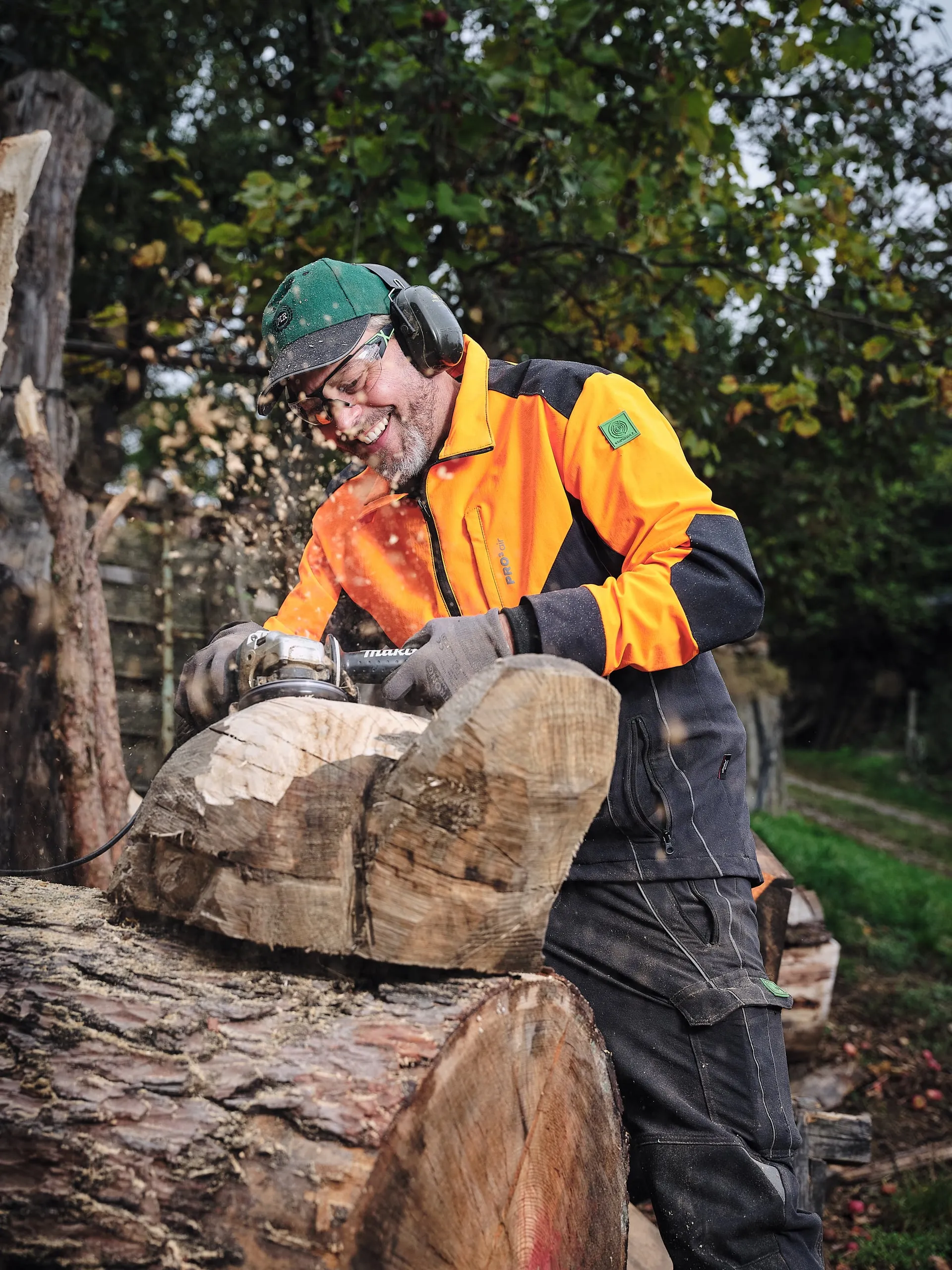Ein Mann in orangefarbener Schutzkleidung bearbeitet im Wald einen Holzstamm.