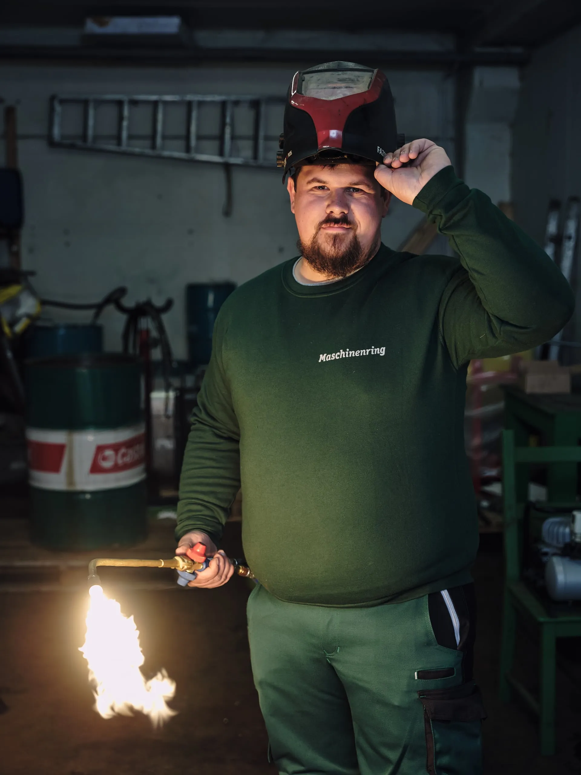 Ein Mann in grünem Pullover und Schutzhelm hebt die Kappe, in der Hand hält er einen Brenner mit Flamme.