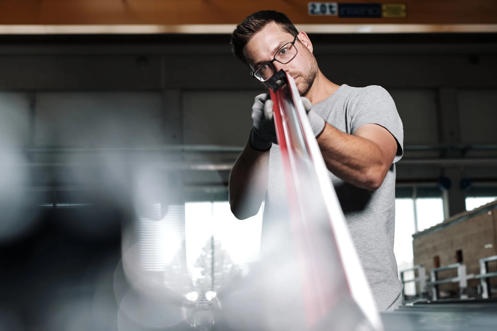 Ein Mann mit Brille und Handschuhen hält in einer Werkstatthalle ein langes, glänzendes Metallprofil vor sich.
