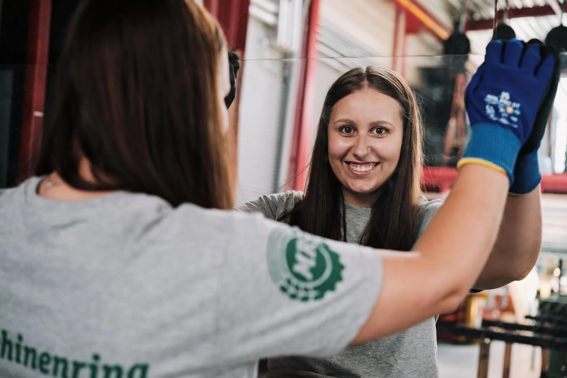 Zwei junge Frauen in grauen Shirts arbeiten in einer Halle, eine hebt lachend die Hand mit blauem Arbeitshandschuh.