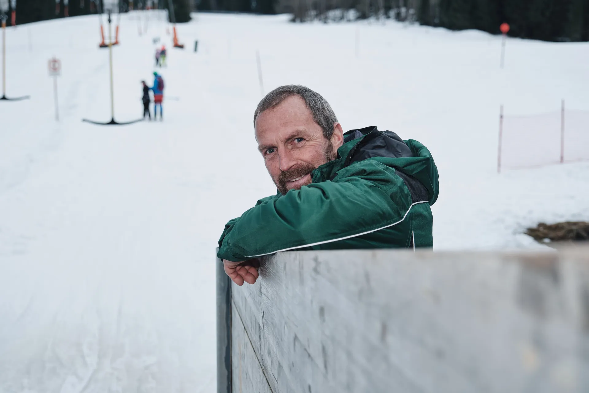 Ein Mann in grüner Jacke lehnt an einer Bande im Schnee, hinter ihm eine verschneite Piste mit Liftanlage.