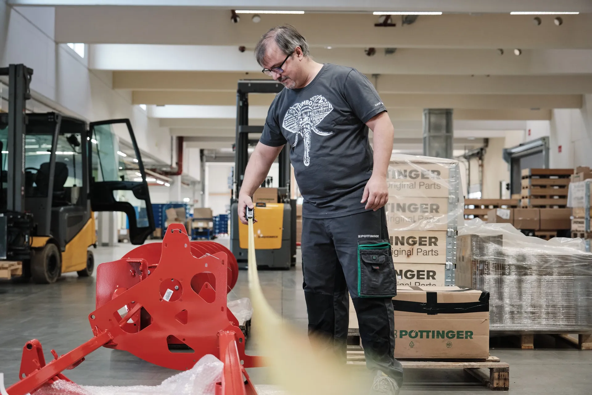 Ein Mitarbeiter zieht ein Maßband entlang eines roten Anbauteils in der Lagerhalle, dahinter ein Stapler.