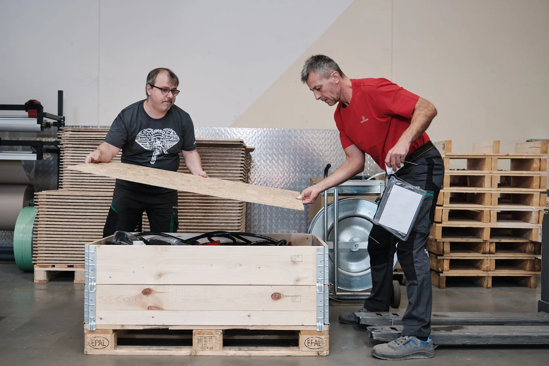 Zwei Mitarbeiter verpacken Teile in einer Holzkiste auf einer Palette in der Versandhalle.
