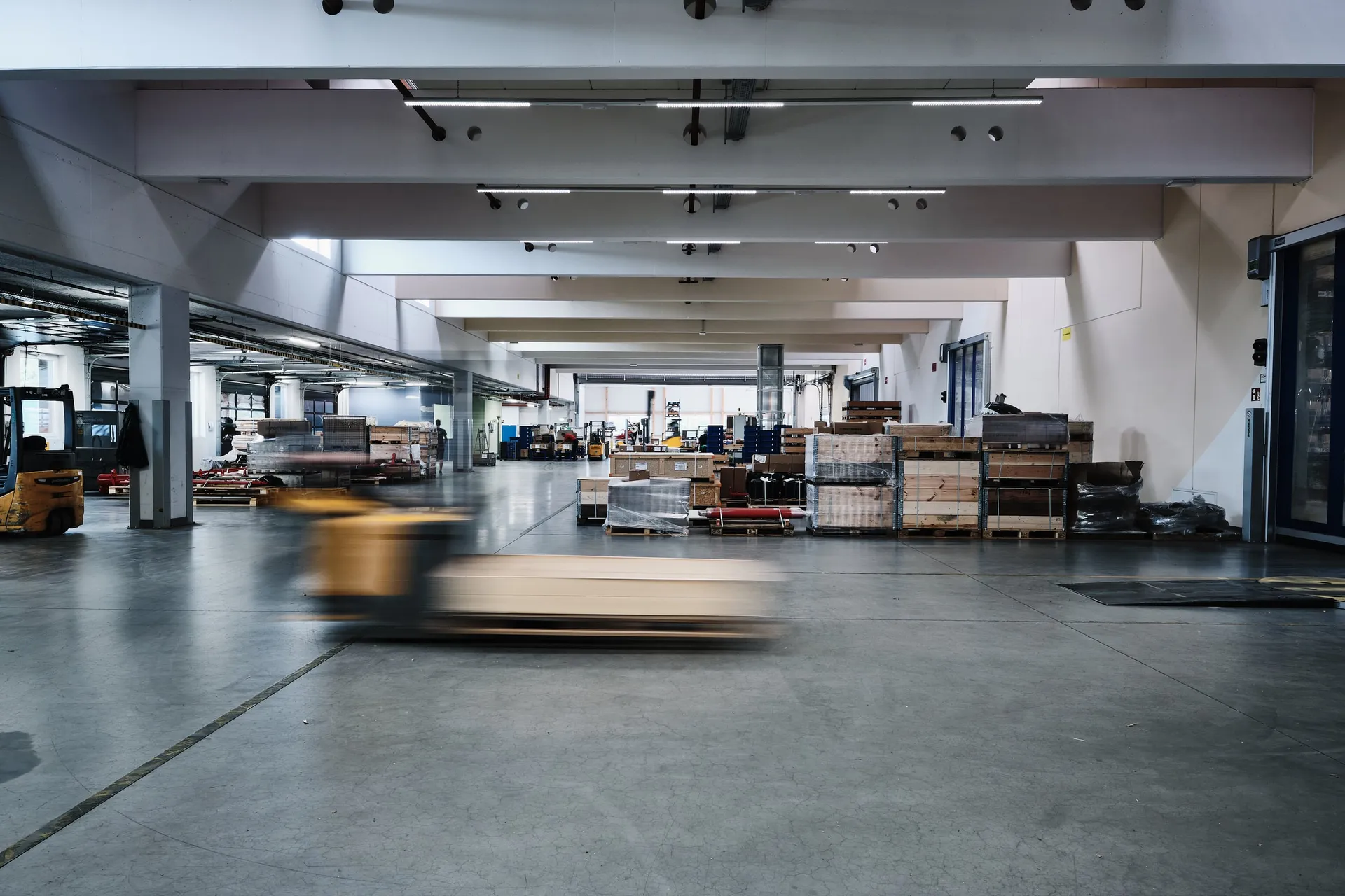 Weite Lagerhalle mit Blick in die Tiefe, im Vordergrund zieht ein Hubwagen in Bewegungsunschärfe vorbei.