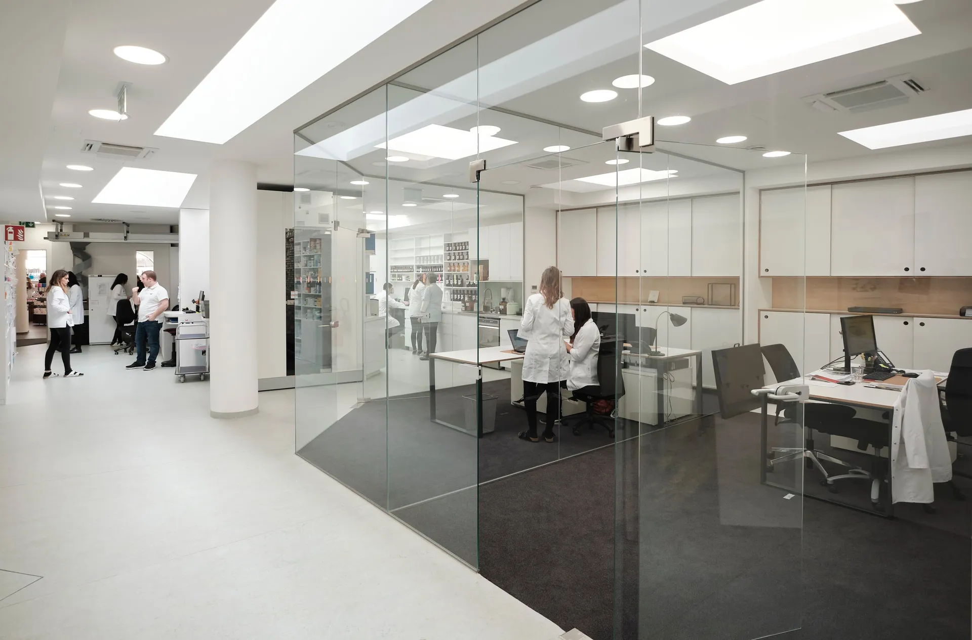 Blick in den hellen Arbeitsbereich einer Apotheke hinter Glaswänden, mehrere Personen arbeiten an Tischen.