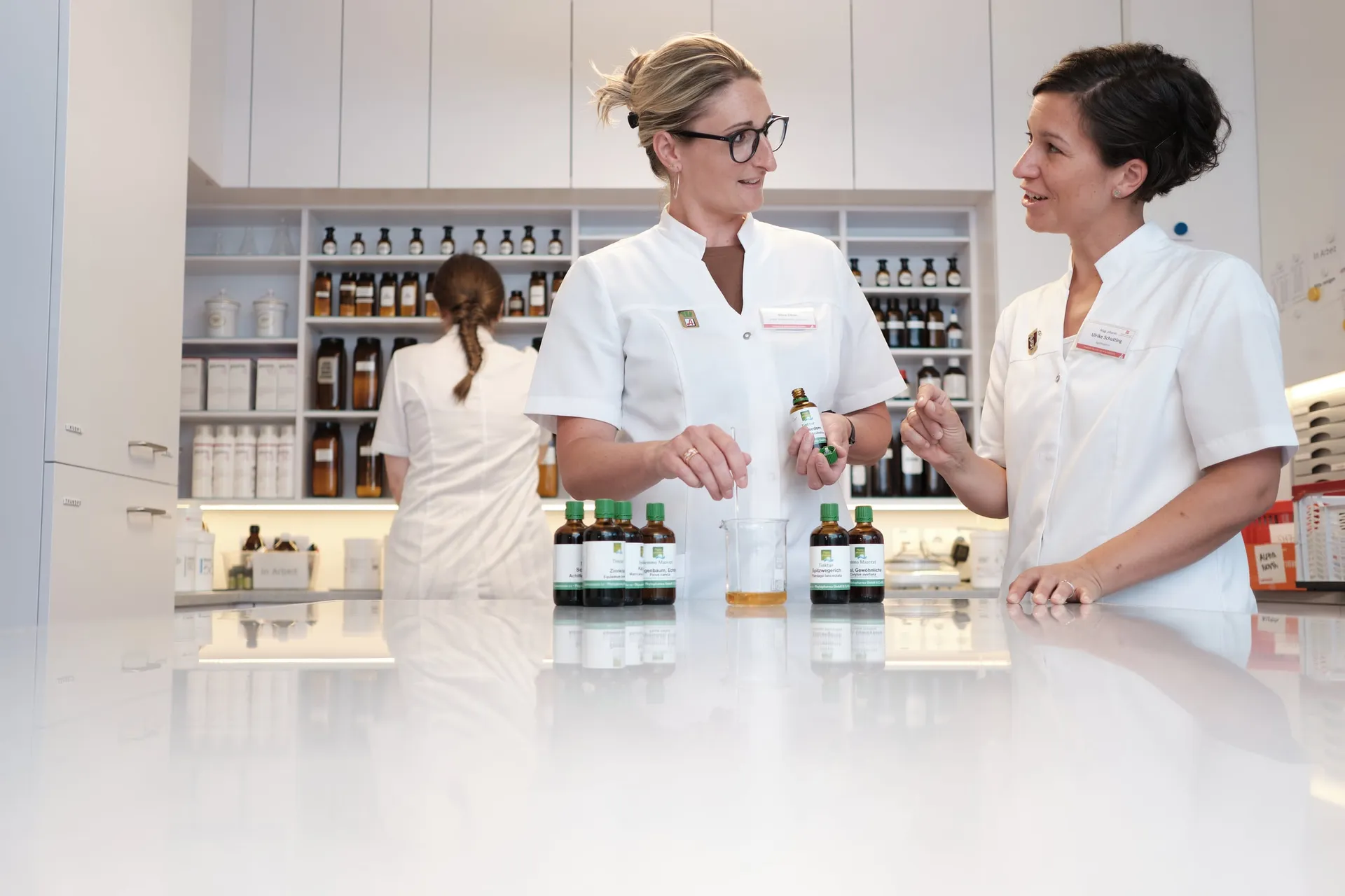 Zwei Apothekerinnen in weißen Kitteln besprechen sich an einer Theke, im Hintergrund arbeitet eine Kollegin vor den Regalen.