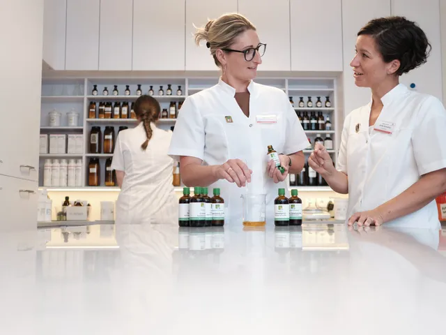Zwei Apothekerinnen in weißen Kitteln besprechen sich an einer Theke, im Hintergrund arbeitet eine Kollegin vor den Regalen.
