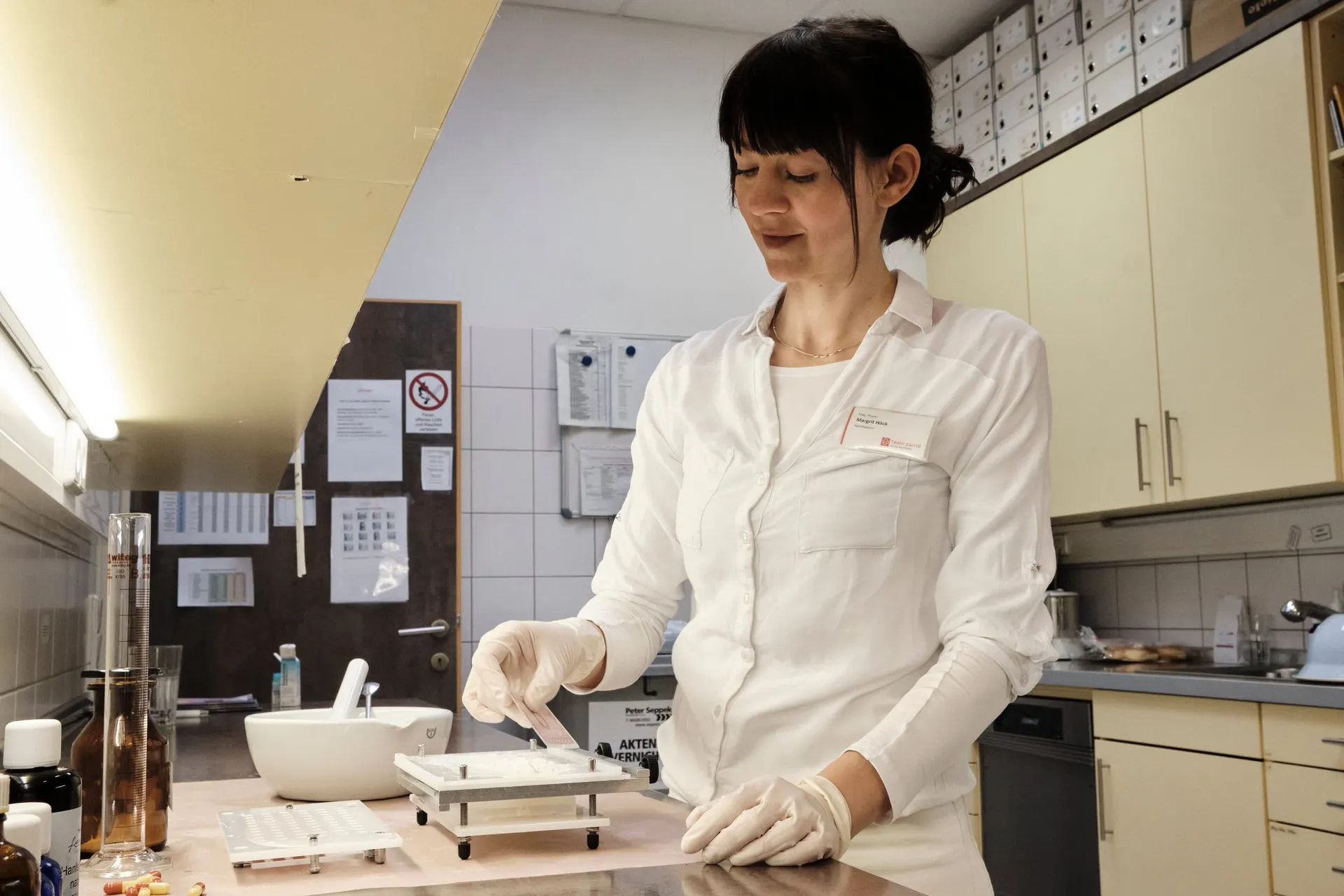 Eine Mitarbeiterin in weißer Bluse und mit Handschuhen stellt an einer Waage eine Rezeptur her, daneben ein Mörser.