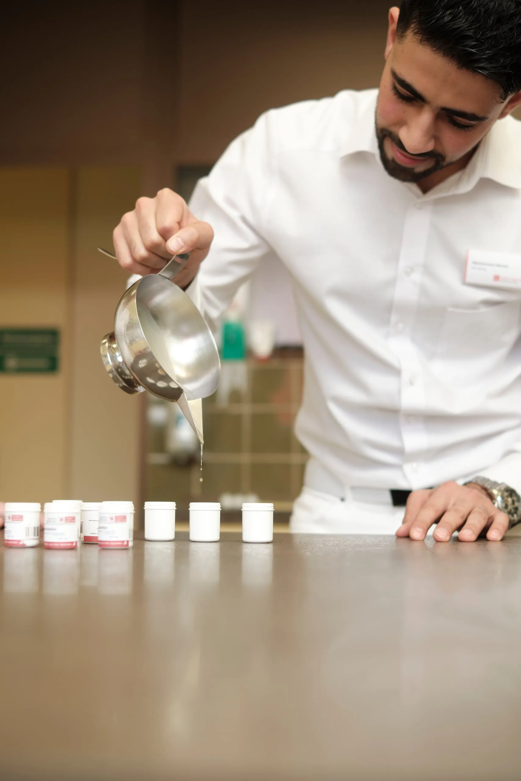 Ein Apotheker in weißem Hemd gießt Flüssigkeit aus einer Metallkanne, davor stehen kleine Döschen.