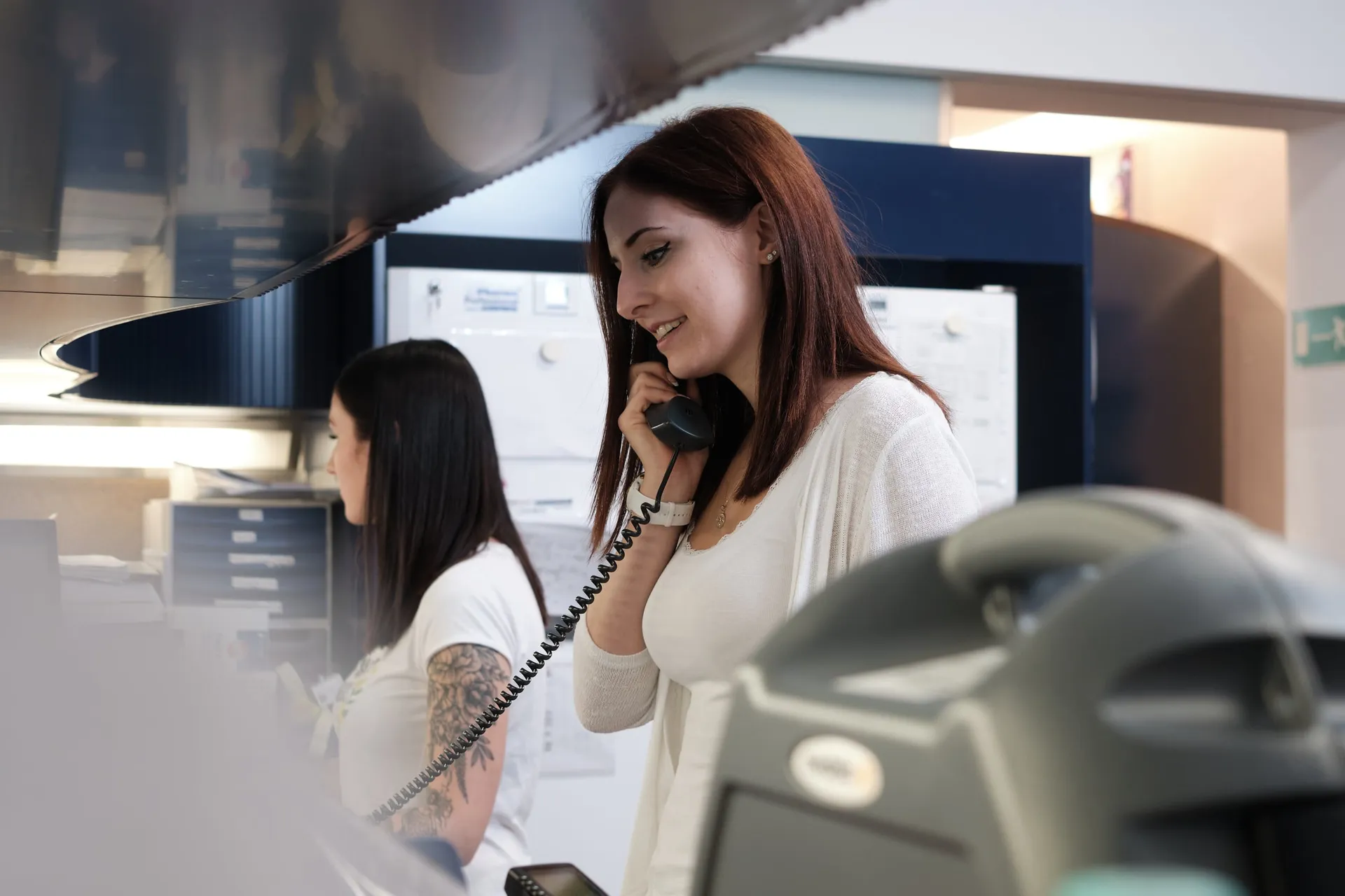 Eine Mitarbeiterin telefoniert, im Vordergrund ein Handscanner, im Hintergrund arbeitet eine Kollegin.
